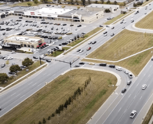 Anayolun hemen yanında büyük bir otopark alanı ve devamında marketlerin yer aldığı alışveriş merkezi; arabalar otoparka girmek için sıra halinde bekliyor.